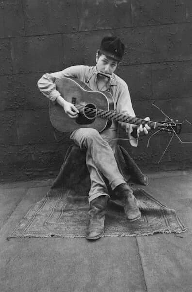 Black and white photograph of a person in work clothes playing an acoustic guitar while seated on a blanket against a brick wall.