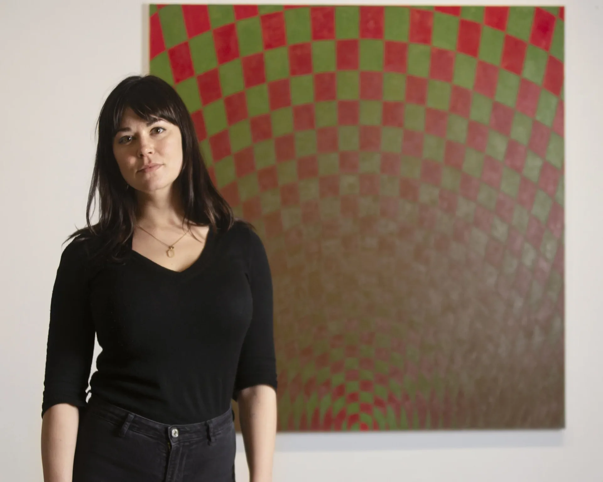 Woman with dark bangs in a black v-neck top stands before a red and green checkerboard-patterned painting at LatchKey Gallery.