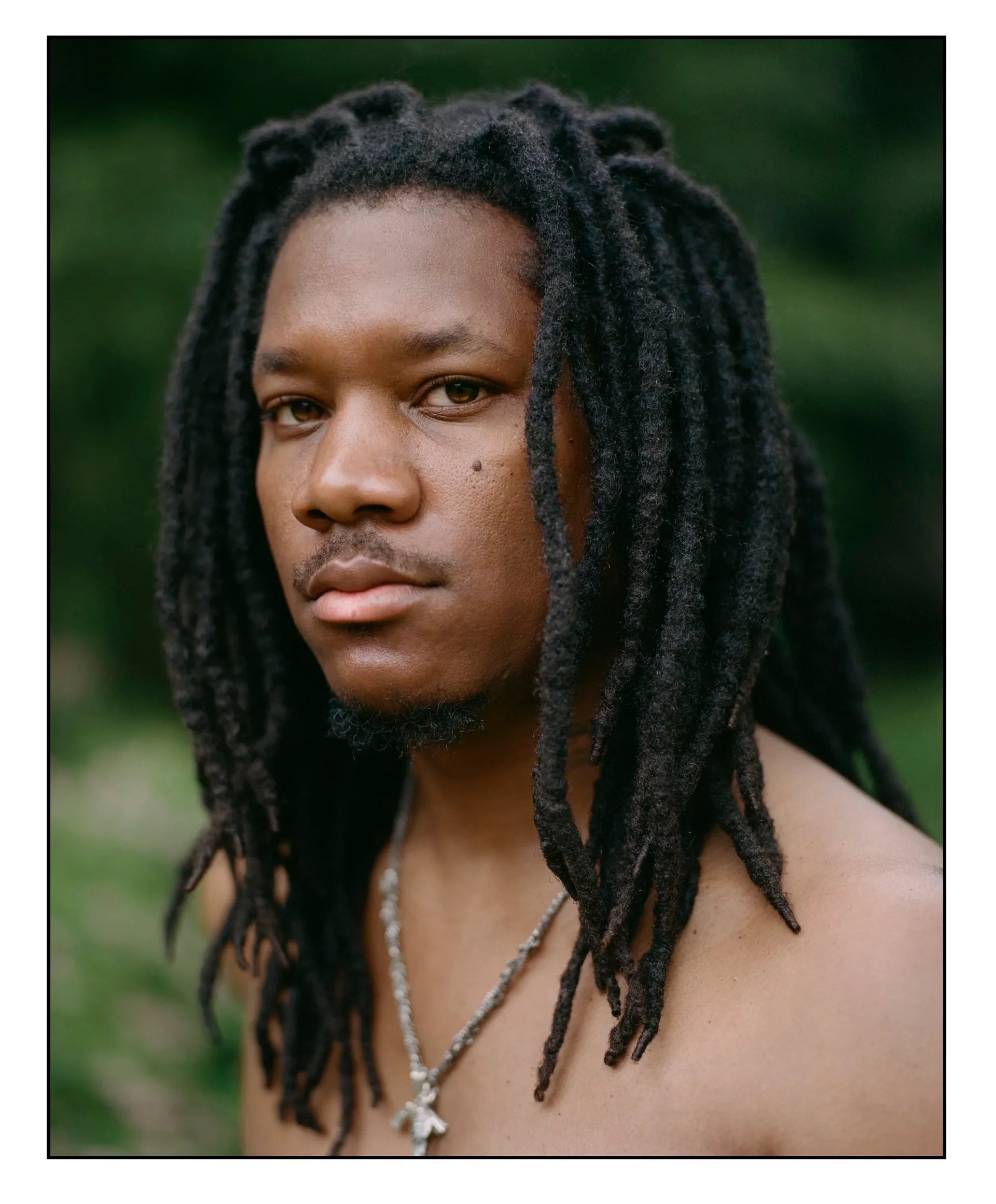 Close-up portrait of a shirtless man with long dark dreadlocks and a beaded necklace against a blurred green outdoor background.