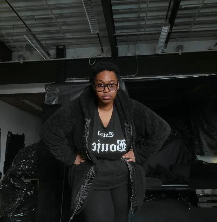 Woman in glasses and a black fuzzy coat over a graphic t-shirt stands with hands on hips in a dim industrial studio space.