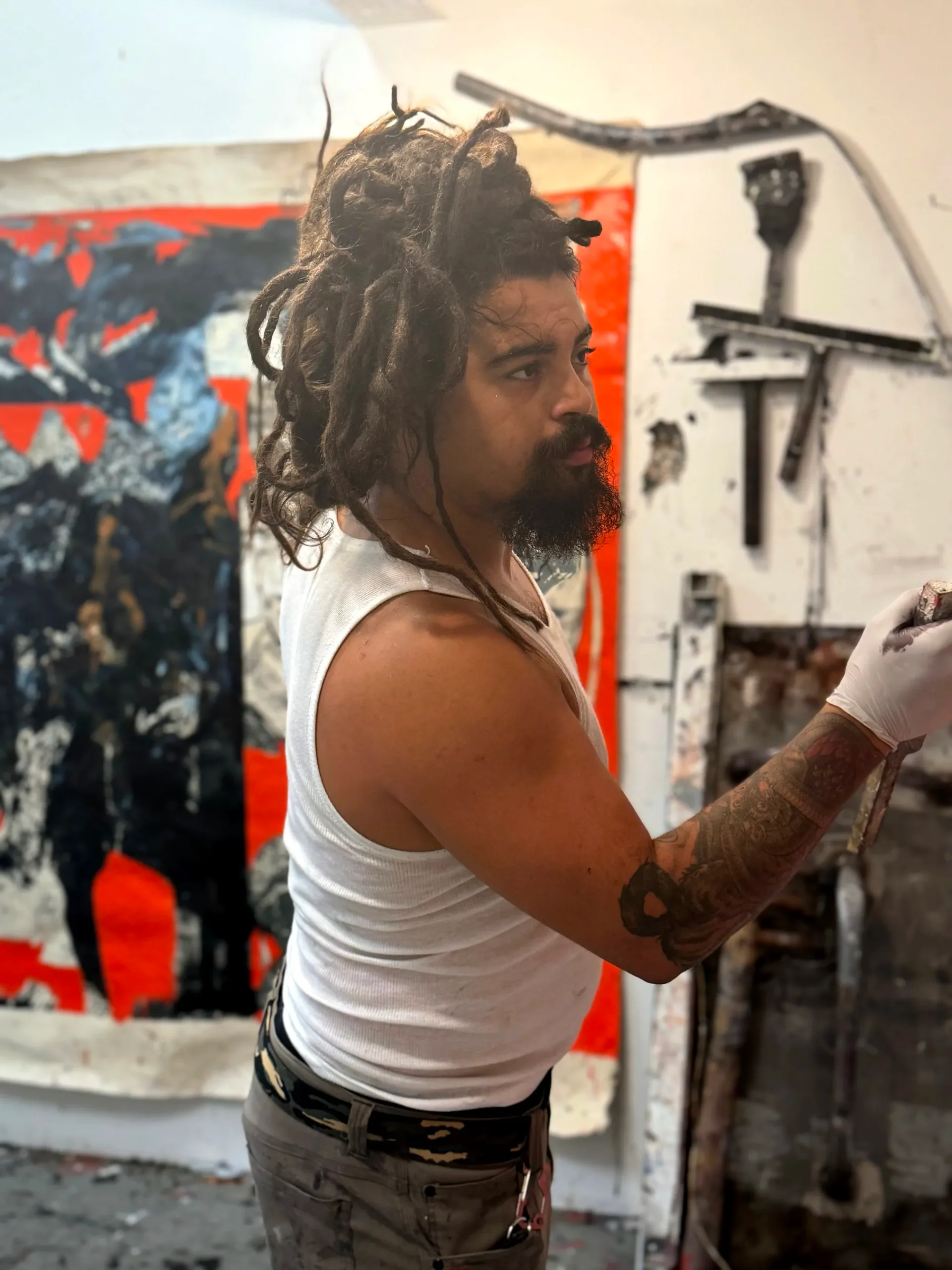 Man with dreadlocks tied up wearing a white tank top works with a gloved hand near tools, red and black artwork visible behind him.