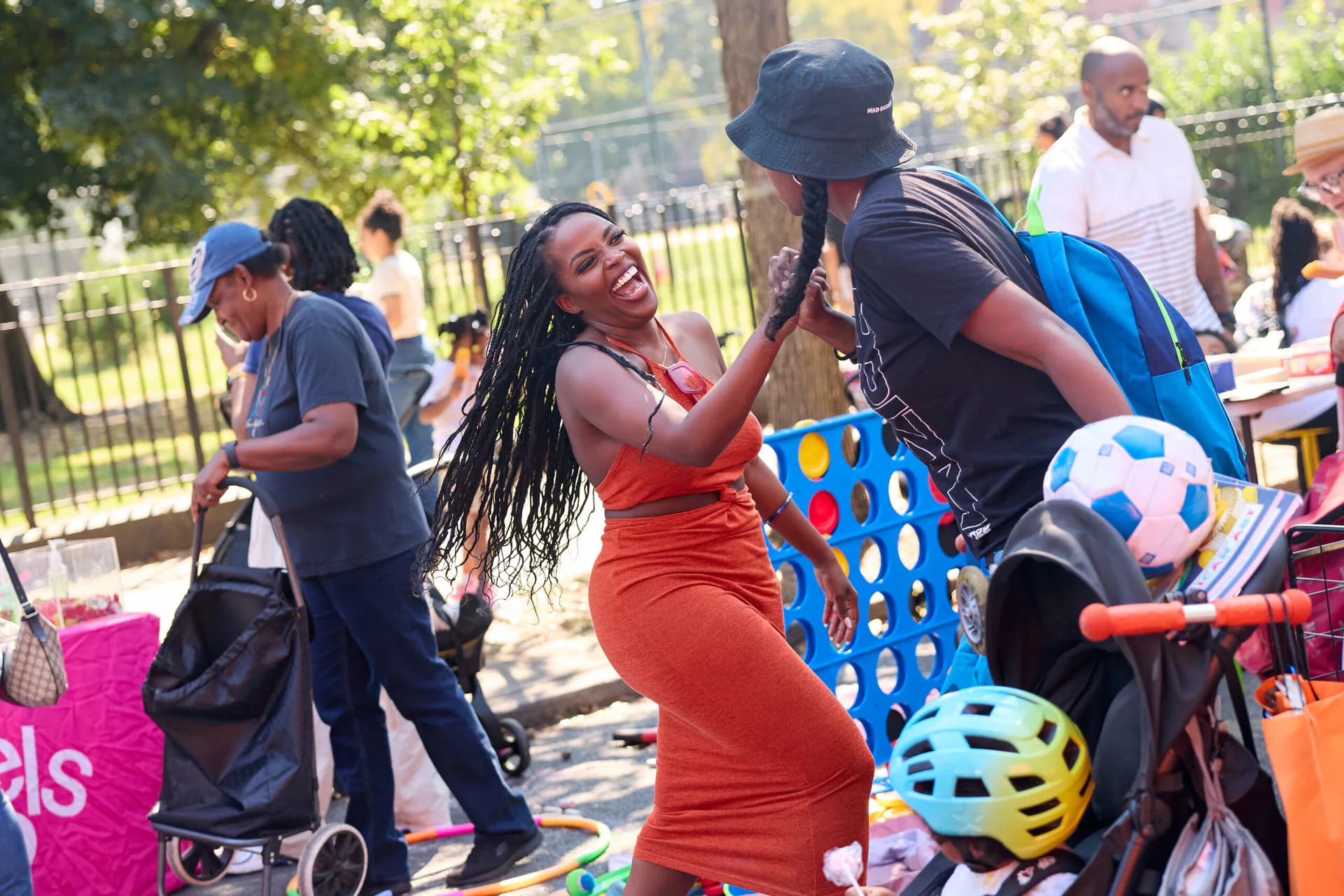 Outdoor community event with people engaged in activities including sports and games; a woman in an orange dress high-fives a person in dark clothing, with colorful balls and park setting visible.