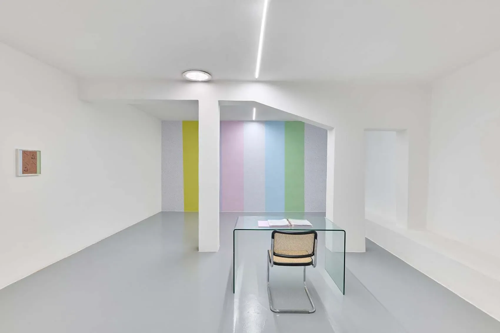 Gallery interior with white walls, vertical colored light panels (yellow, lavender, pink, blue, green), white column, and sculptural furniture pieces on polished floor.