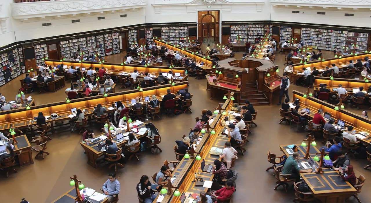 Overhead view of a library reading room with curved wooden study desks arranged in rows, patrons seated and studying, surrounded by dark wood shelving and cream-colored walls.