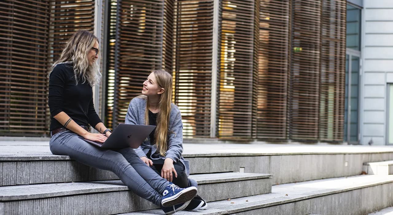 Two young women sitting on outdoor steps in conversation, wearing casual clothing, with a modern metal shutter/gate visible in the background.