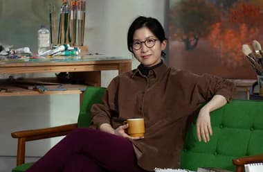 Person wearing glasses and a brown shirt sitting on a green couch, holding a yellow mug, in a studio setting with art supplies and canvases visible.