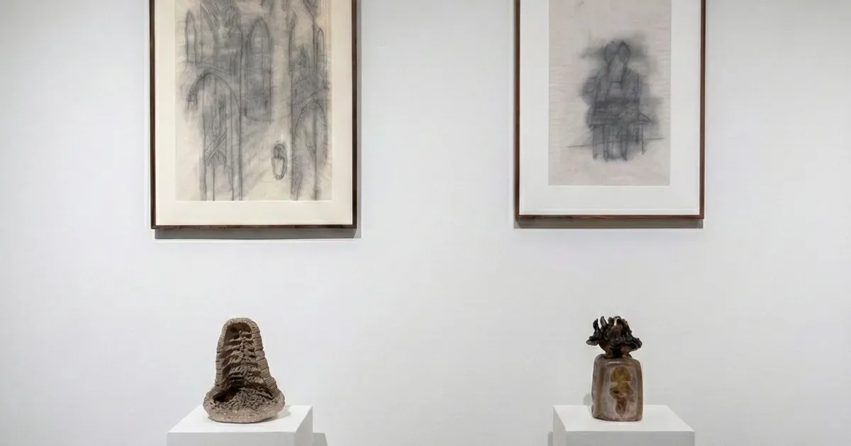 Gallery wall displaying two framed drawings above two sculptural vessels on pedestals, minimalist presentation on white wall.