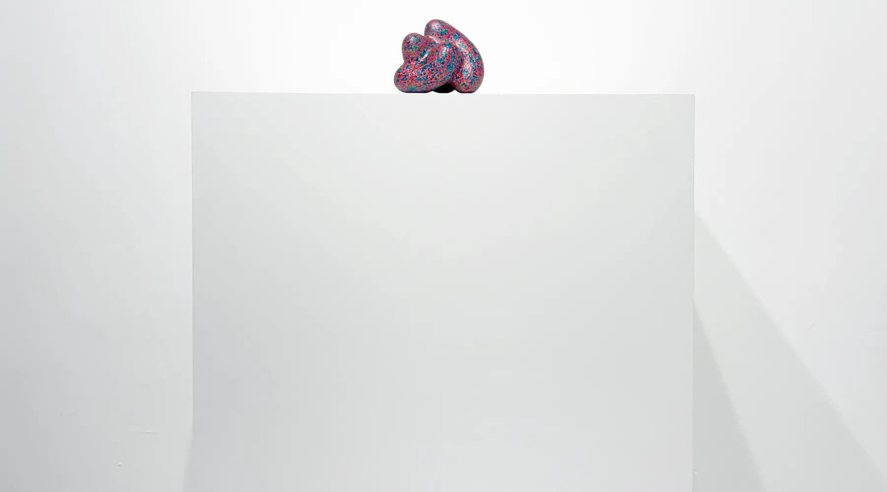 Small purple-pink sculptural object sits atop white architectural form against minimalist white background.