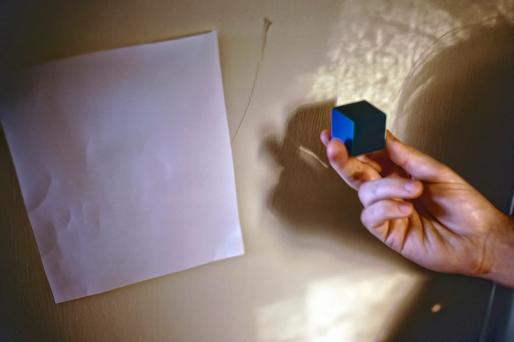 Hand holding a small geometric black and blue cube object, casting a shadow on white paper in warm natural light.