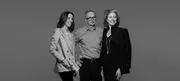 Black and white portrait photograph of three people standing against a neutral gray background.