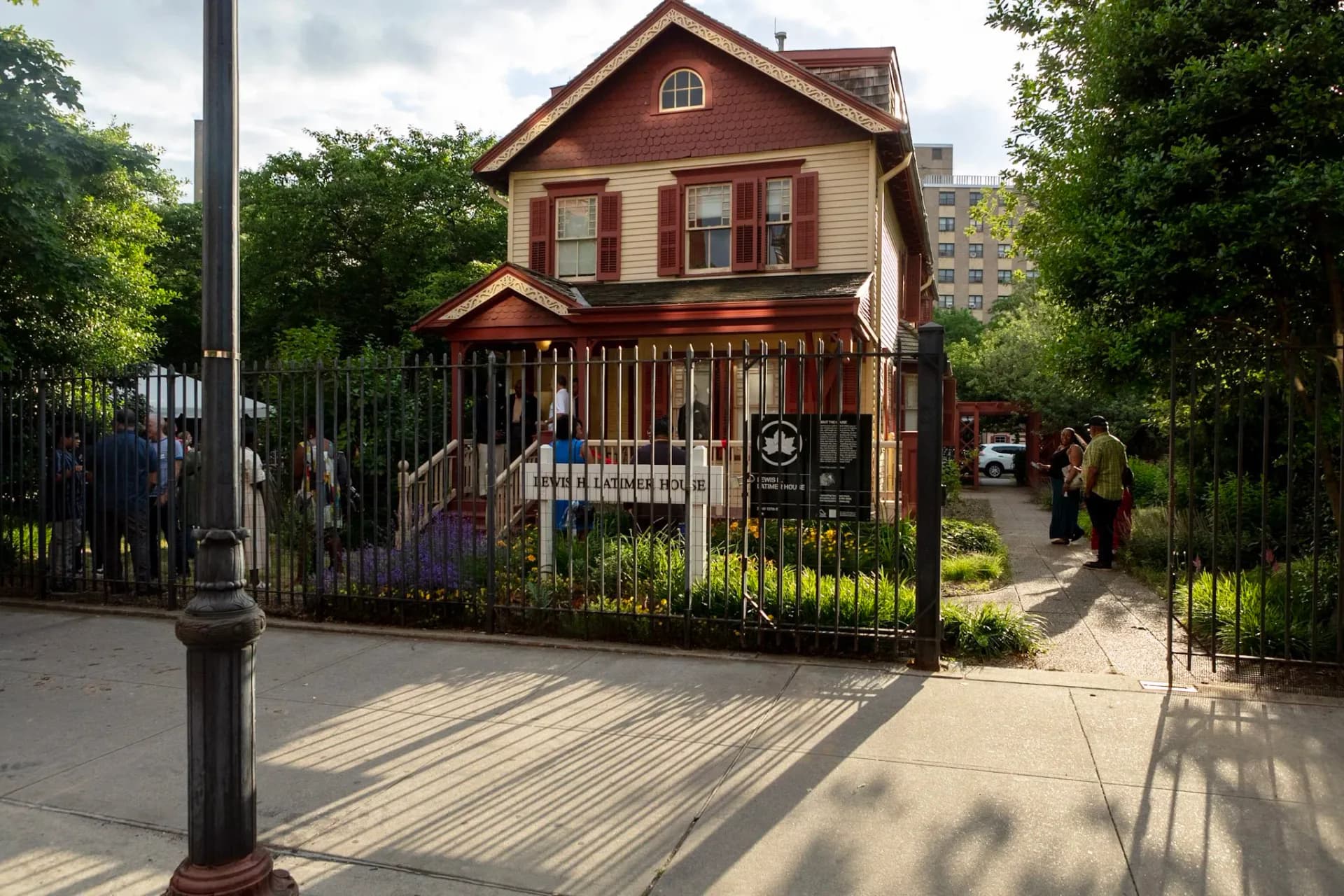 Red brick two-story house with white trim and metal fence, surrounded by green foliage and trees on an urban street.