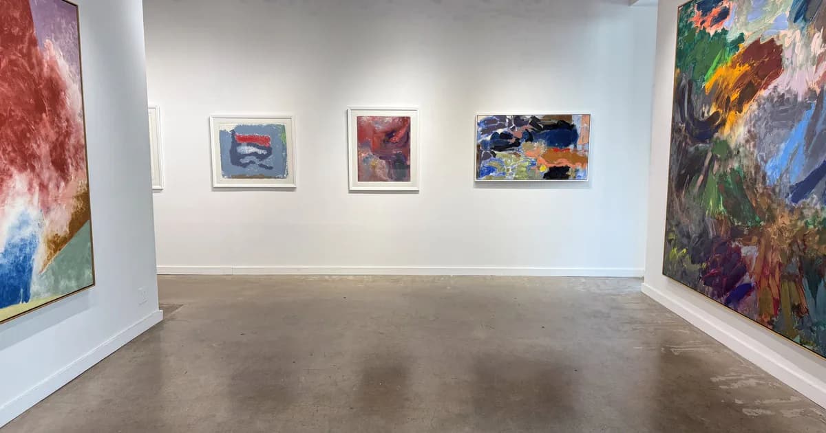 Gallery interior with white walls displaying five framed artworks of varying sizes, polished concrete floor, and large colorful abstract paintings on side walls.