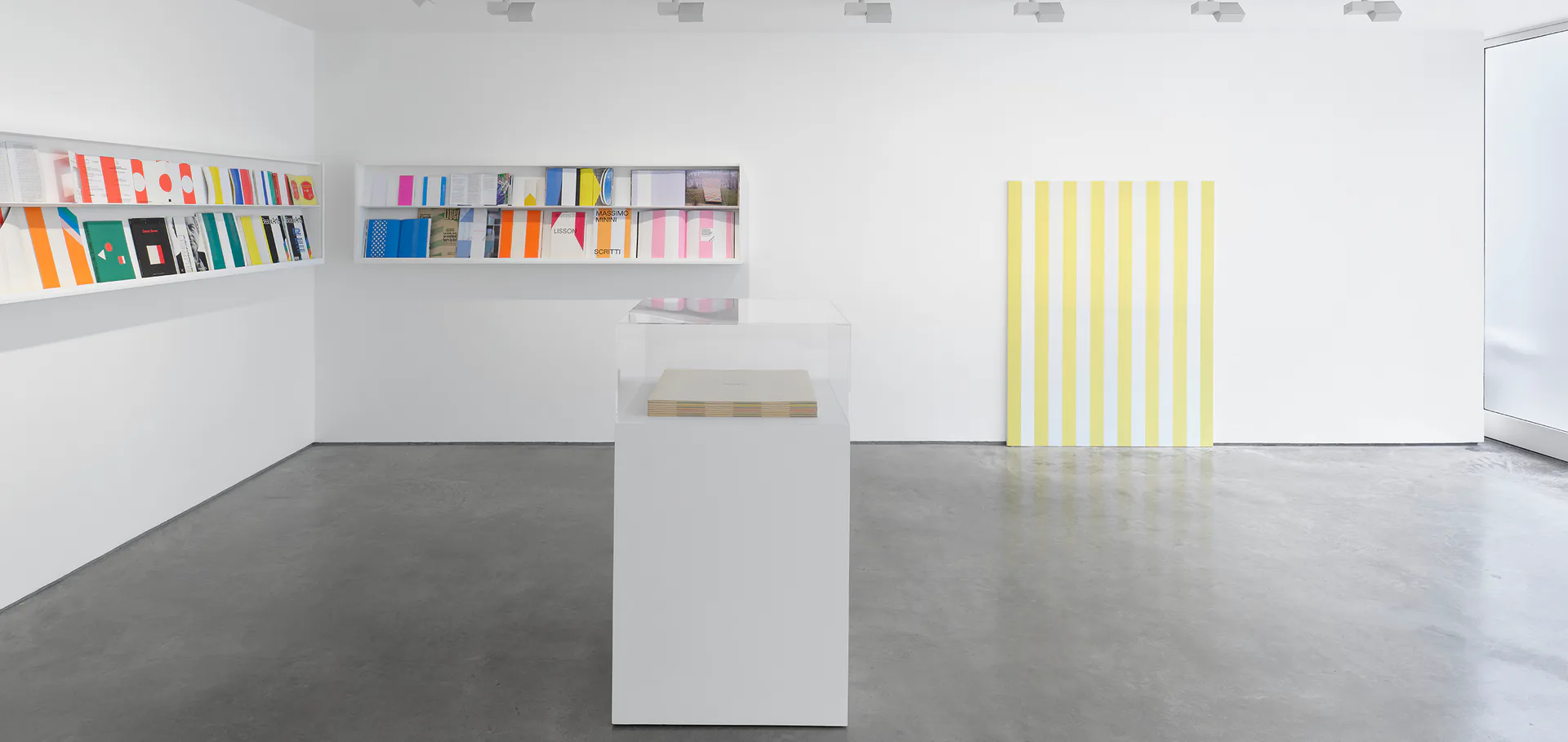 Lisson Gallery exhibition space with white walls, polished concrete floor, featuring colorful books on wall-mounted shelf, central white pedestal, and yellow vertical striped panel.