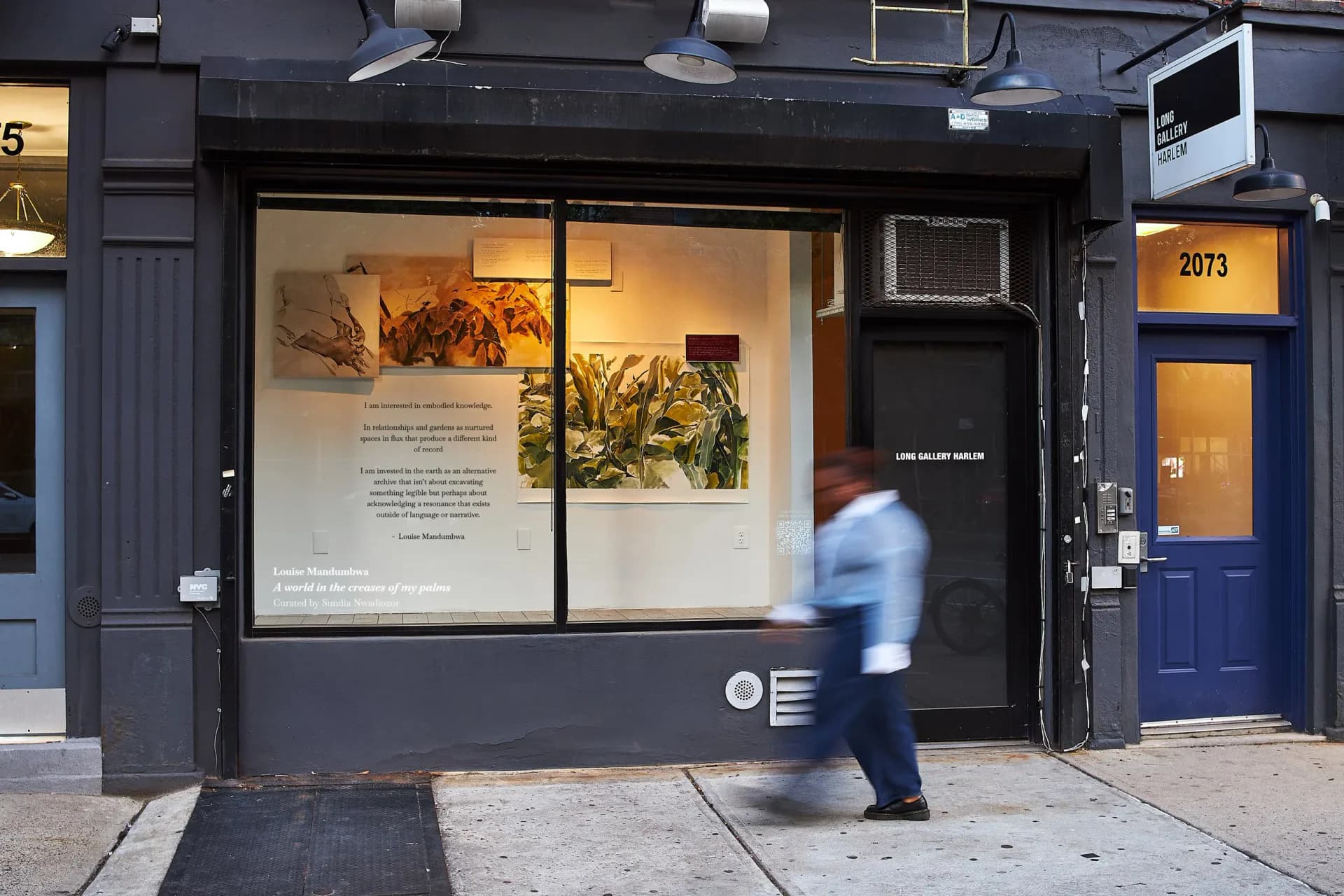 Long Gallery Harlem storefront exterior with black facade, large window displaying artwork, blue door, pedestrian passing by.