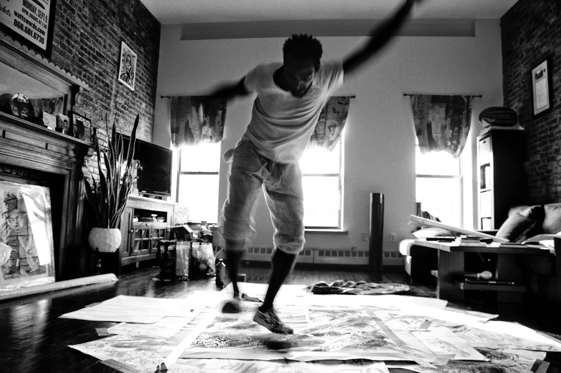 Black and white photograph of artist in motion painting on large floor canvas in studio with brick walls and windows.
