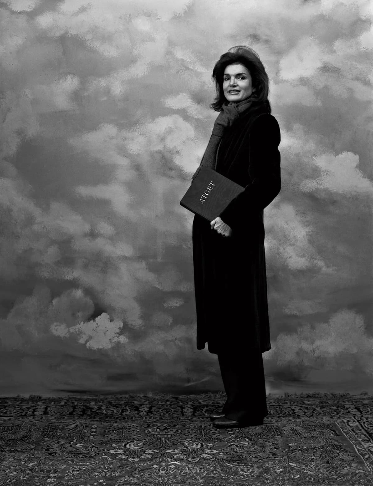 Black and white portrait of a woman in dark formal clothing holding a book, standing against a dramatic cloudy sky backdrop.