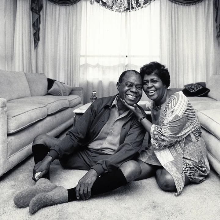 Black and white photograph of a man and woman smiling together on a carpeted floor with a sofa and curtains behind them.