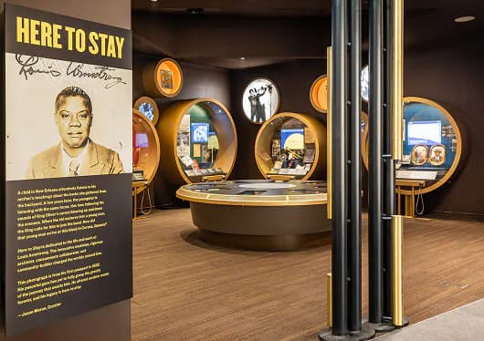 Interior photograph of a museum gallery space with dark walls, gold-rimmed circular displays, and a sign reading 'HERE TO STAY' on the left.