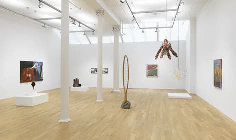 Bright gallery interior with wooden floors, white walls, exposed columns, and various artworks displayed including sculptures and paintings.
