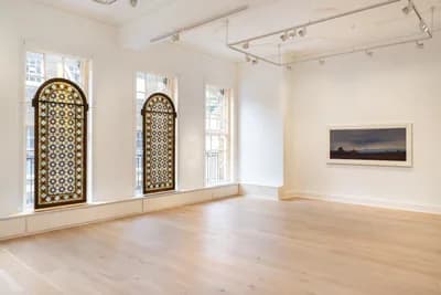 Gallery interior with light wood floors, two ornate arched doorways with geometric patterns, white walls, and a dark landscape painting on the right.