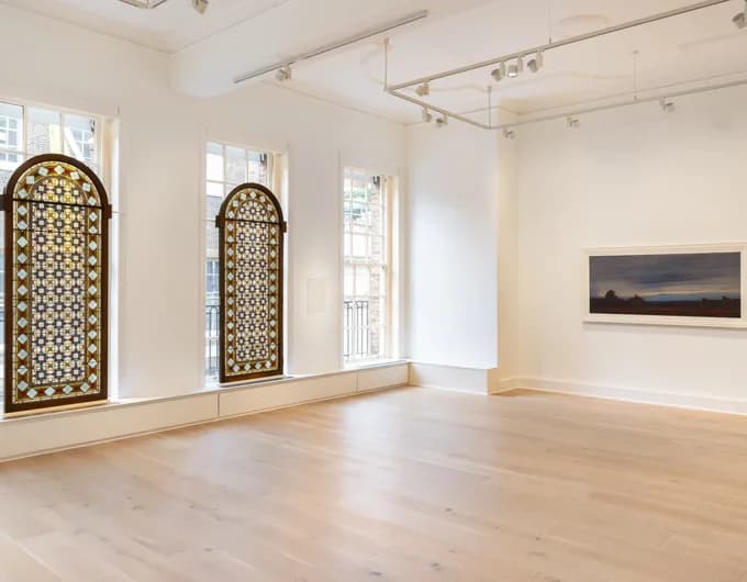 Minimalist gallery space featuring polished wood floors, two decorative arched mirrors or panels with geometric gold patterns, and a dark framed artwork on pale wall.
