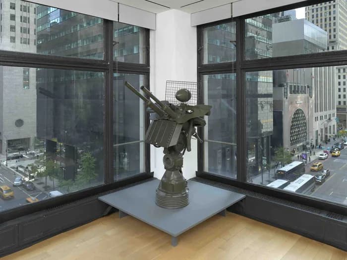 Modern gallery corner with expansive floor-to-ceiling windows overlooking urban cityscape, blonde wood floor, and bronze sculptural figure on blue pedestal.