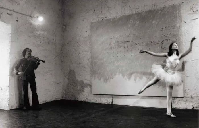 Black and white photograph or video still showing musician with violin and ballet dancer in motion against weathered concrete wall in industrial space.