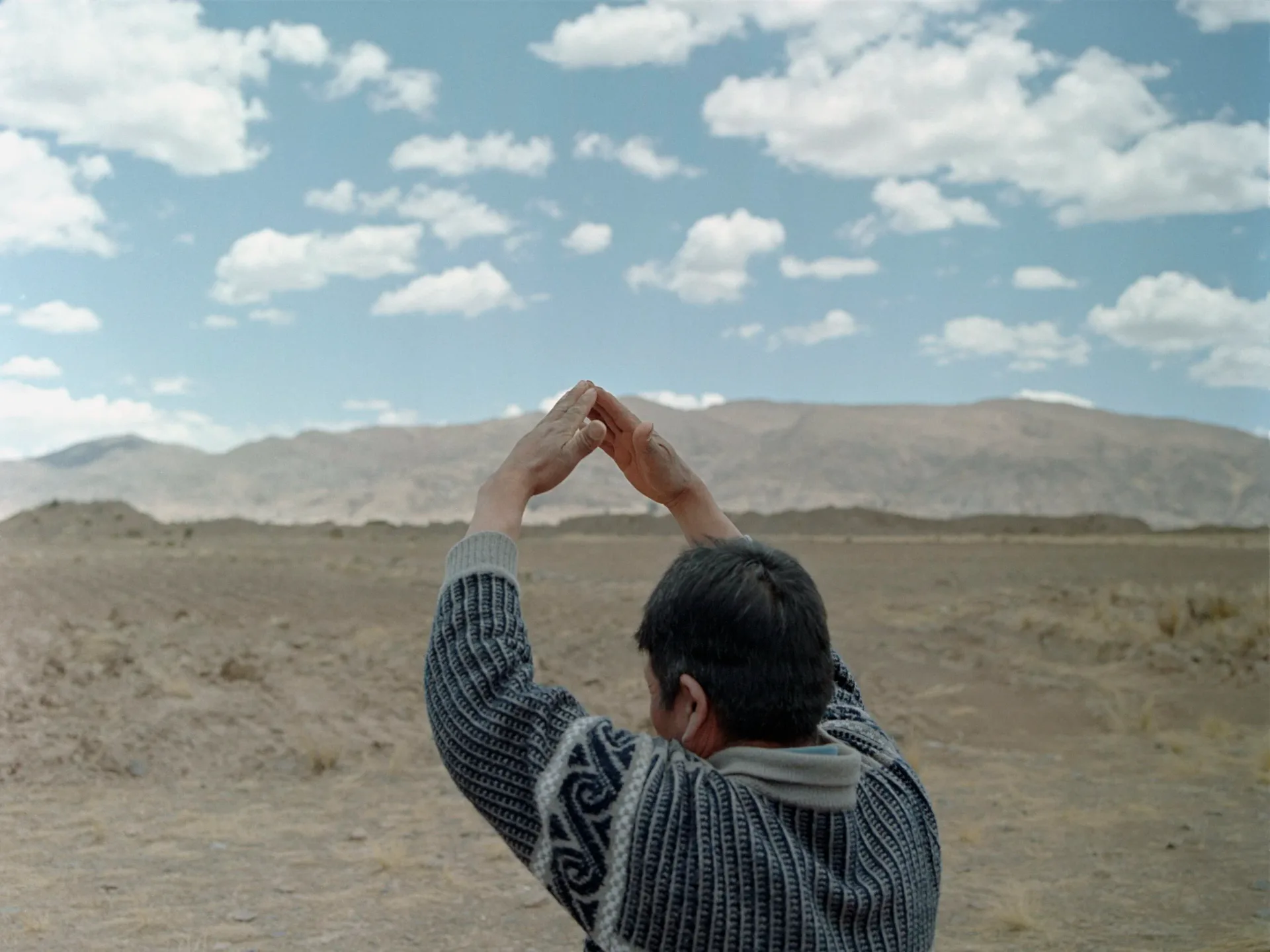 Person from behind forming a heart shape with their hands toward distant mountains under blue sky with white clouds.
