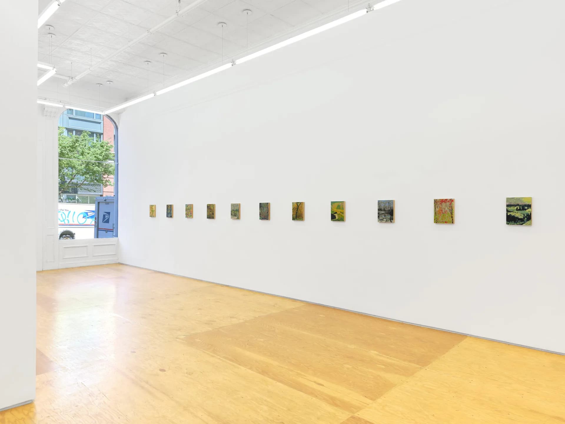 Gallery interior with wooden floor, white walls, and a row of small framed artworks hung at eye level; blue signage visible on left wall.