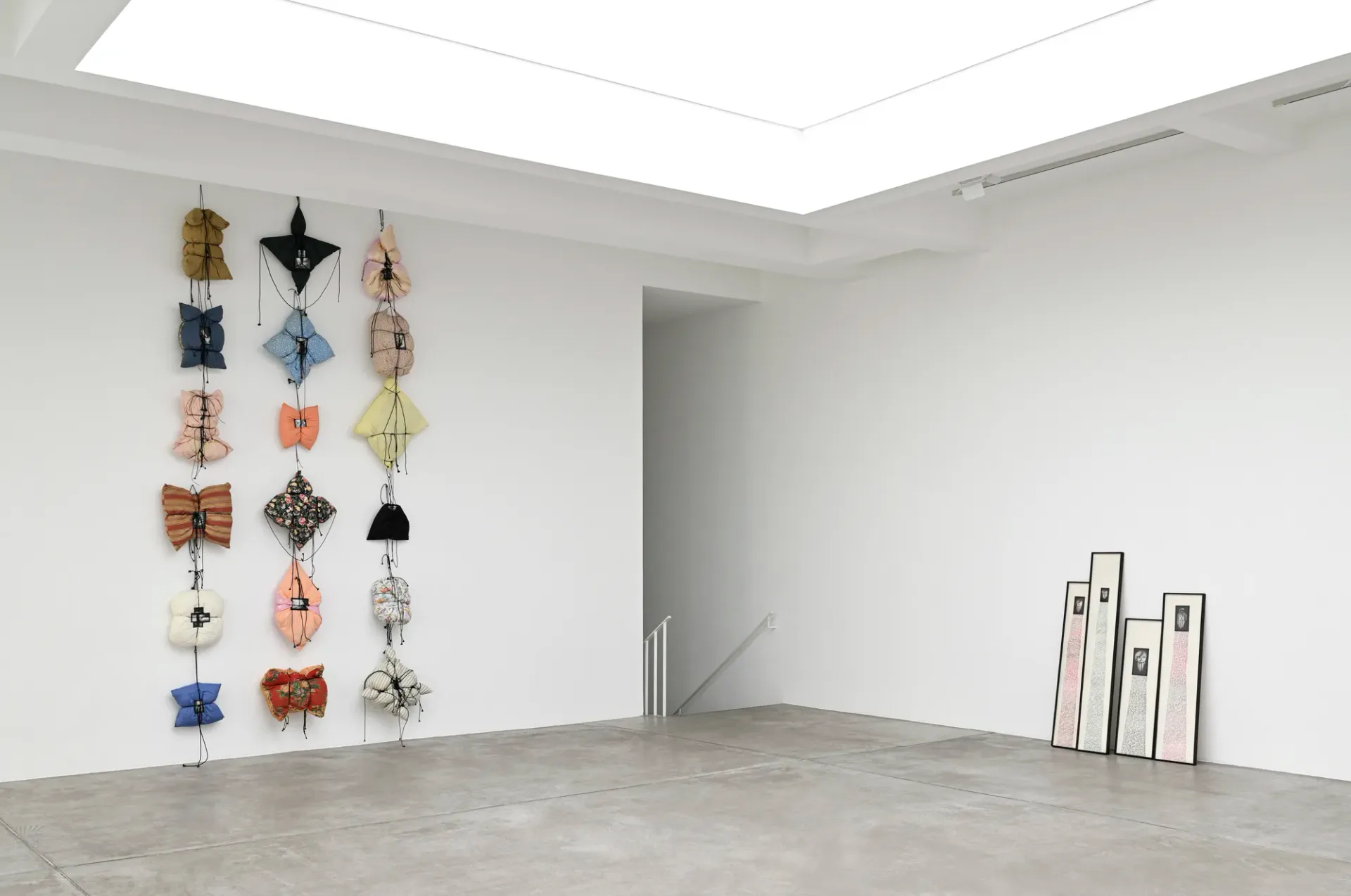 Gallery interior with white walls, skylight, polished concrete floor, and textile artworks arranged vertically on left wall.