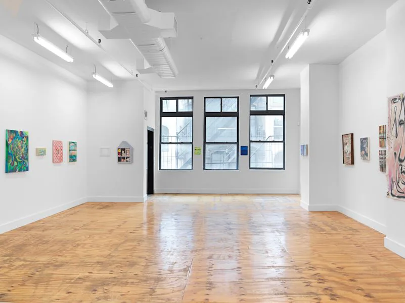Bright, minimalist gallery interior with blonde wood floors, white walls, large windows, and artworks displayed sparsely on walls.