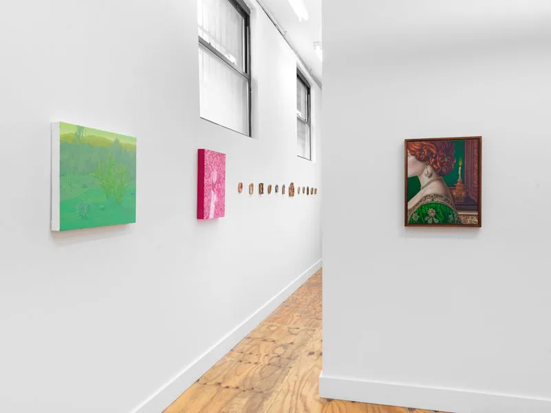 Gallery hallway with windows, wood floors, and small artworks including a green canvas, pink canvas, and patterned mixed-media piece on white walls.