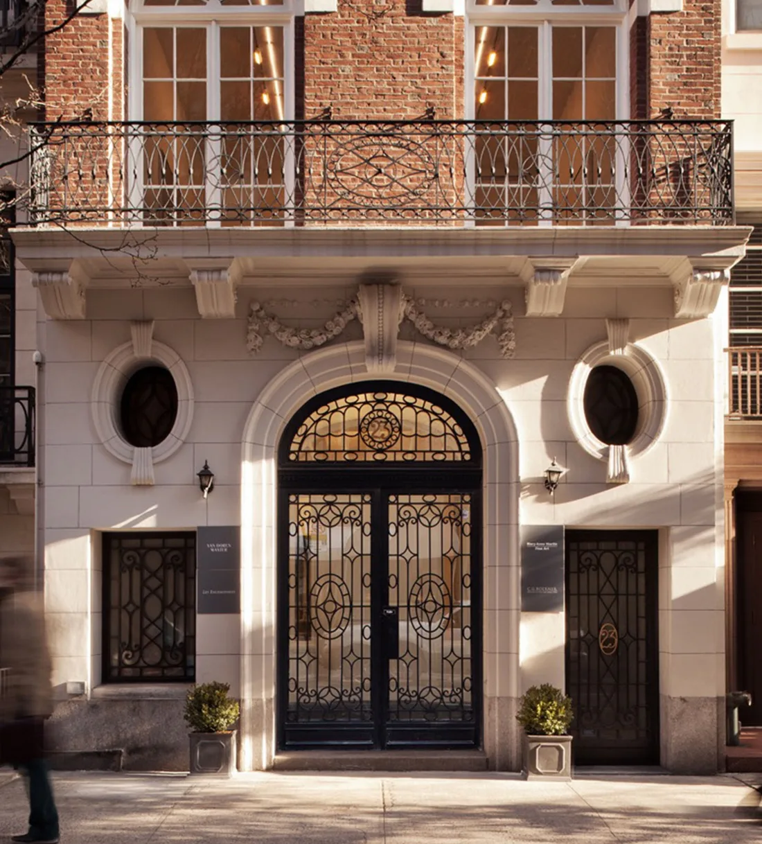 Exterior facade of a Manhattan building with ornate ironwork double doors, oval windows, and a brick balcony above, marking the gallery entrance