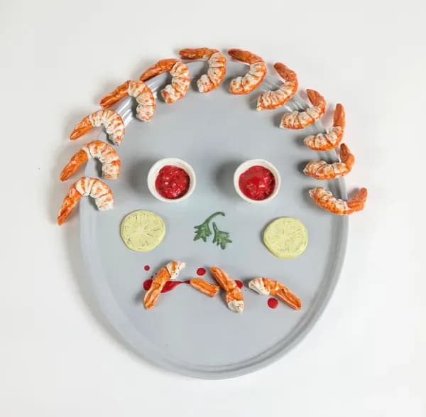 Food art installation: a face composed of shrimp hair, lime slices, tomato sauce eyes, herbs, and red pepper mouth on gray plate.