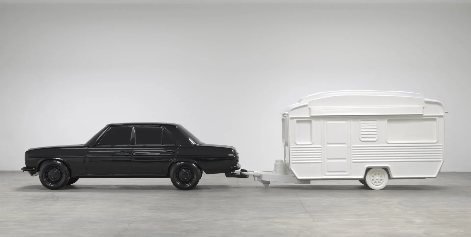 Miniature black sedan car and white travel trailer on gray surface, rendered as a small-scale sculptural work.