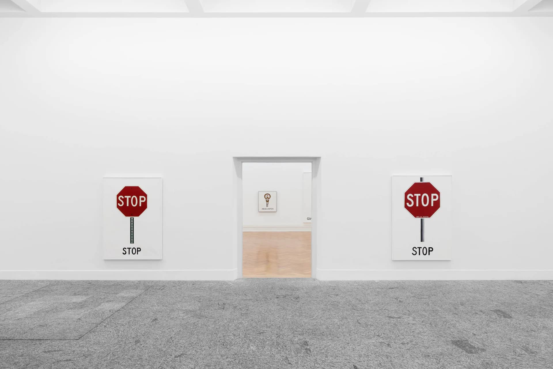 Minimalist installation with two red STOP signs flanking a central gray cabinet with light fixture on concrete floor.