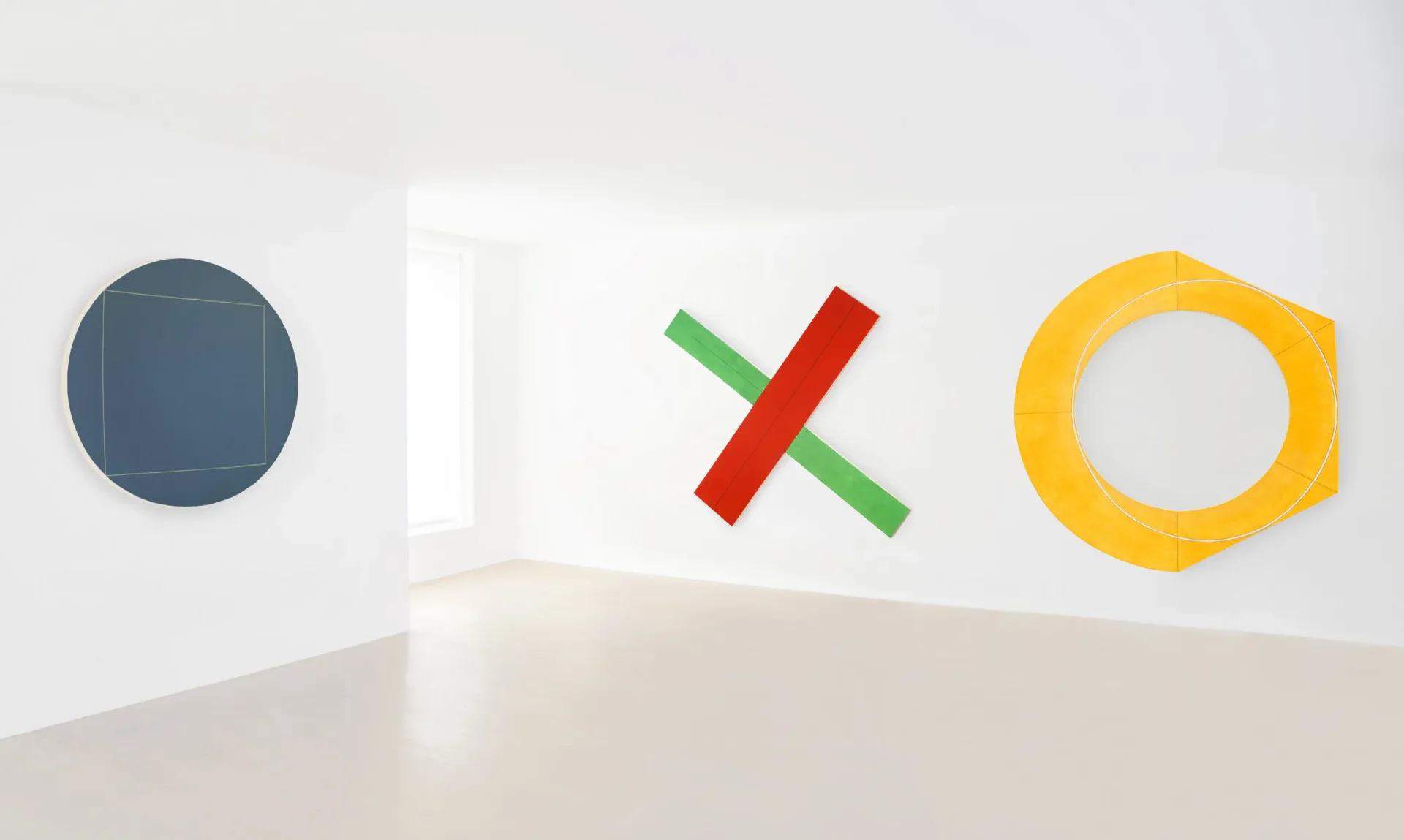 Three-dimensional sculptural objects in a white gallery space: dark blue circle, white rectangular form, red and green crossed bars, and yellow hexagonal ring.