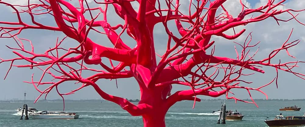 Vibrant red sculptural tree with bare branching limbs installed in shallow water with boats visible in background, likely by Ai Weiwei.
