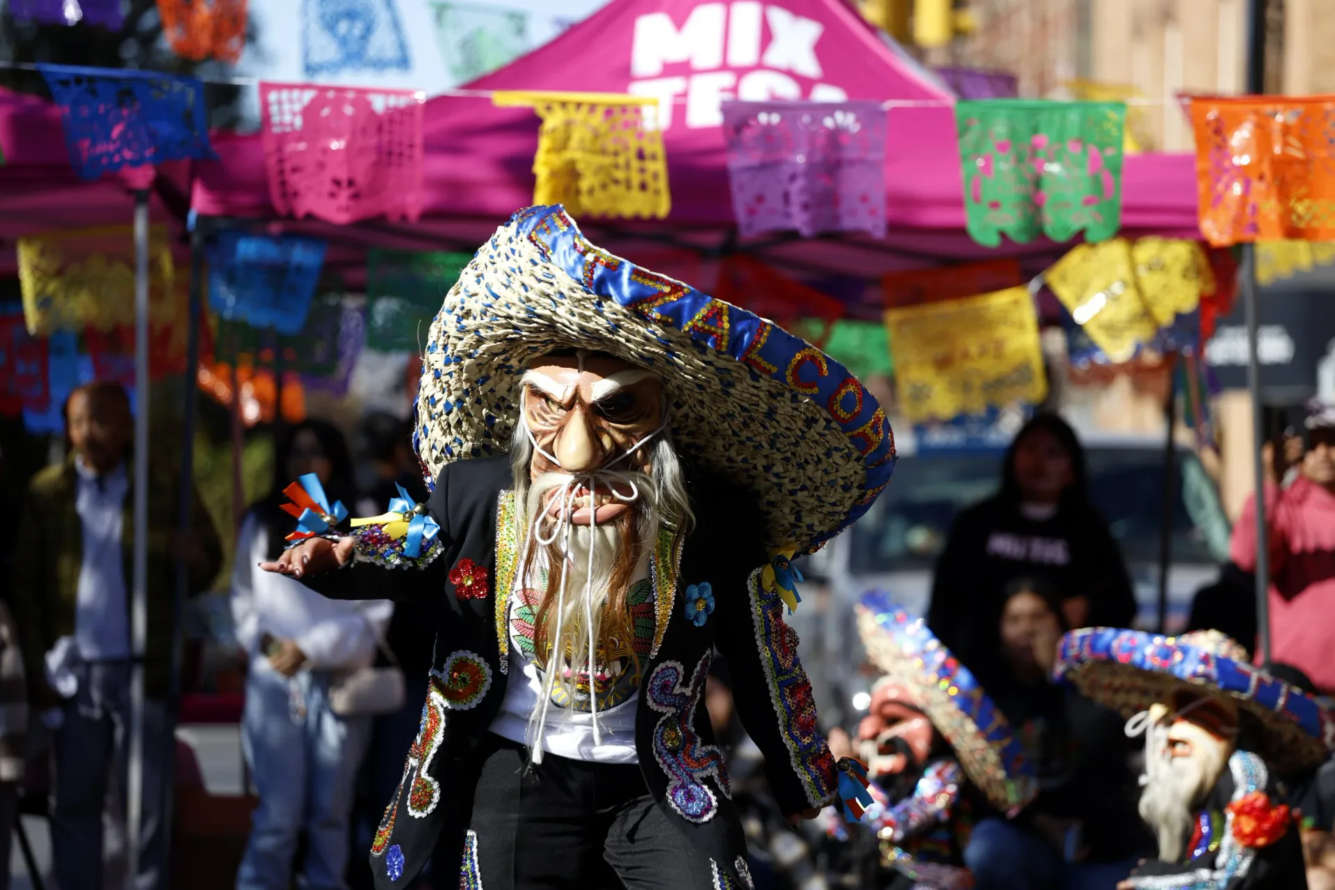 Person in elaborate costume with large woven hat, skeleton-like face paint, and ornate black clothing adorned with decorative elements, at an outdoor festival with colorful papel picado banners.