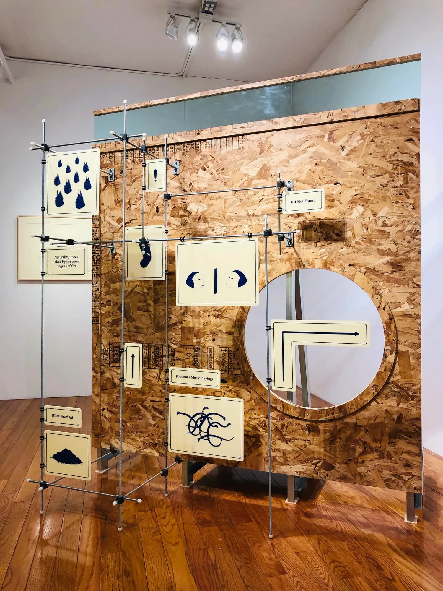 Gallery installation view featuring a large cork-surfaced panel mounted on metal framework with various artworks, drawings, and objects arranged on its surface.