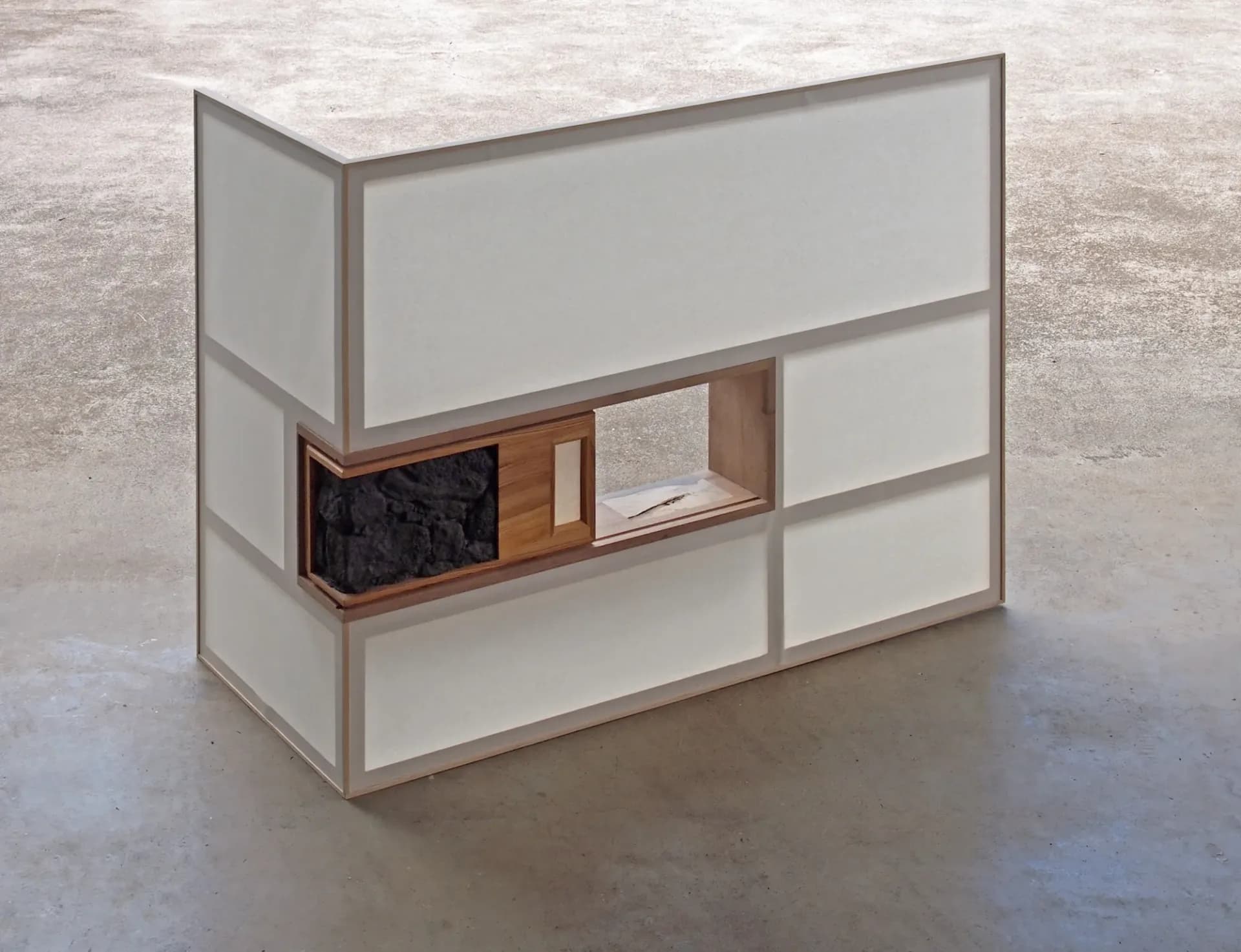Minimalist three-dimensional shelving unit with white panels and a central wooden compartment containing dark and light materials, photographed against neutral background.