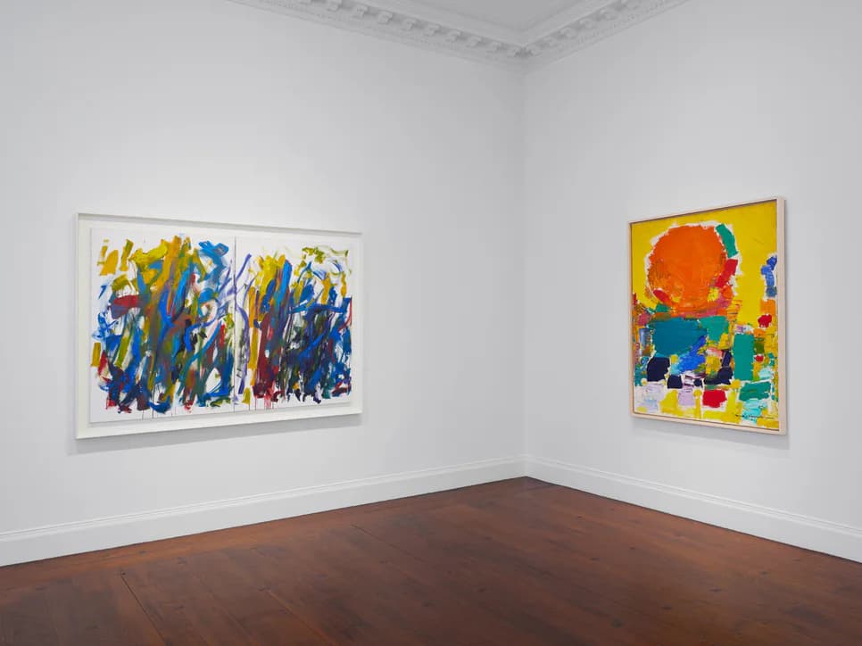 Gallery corner displaying two colorful abstract paintings—one blue and yellow striped, one yellow with orange sun motif.