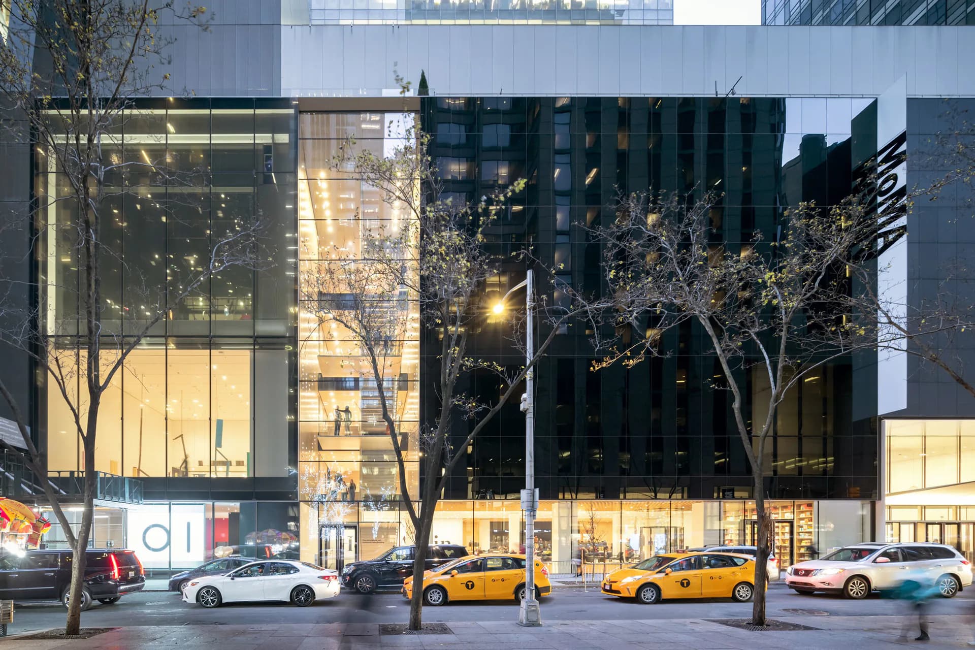 Modern glass and steel building facade at dusk with illuminated interior, bare tree, and yellow taxi cabs on city street.
