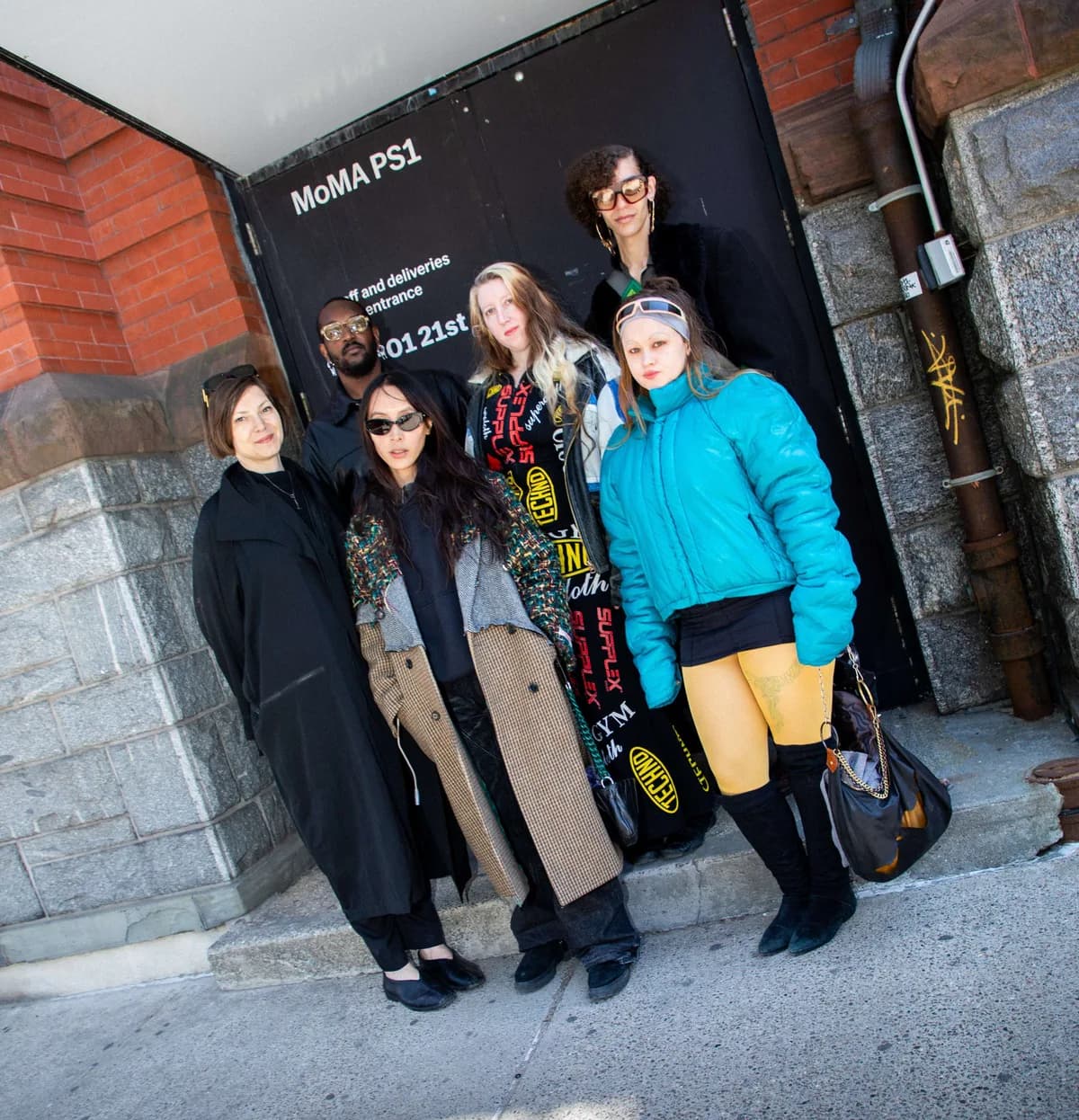 Five people posing together against a dark MoMA PS1 branded wall or sign outdoors.