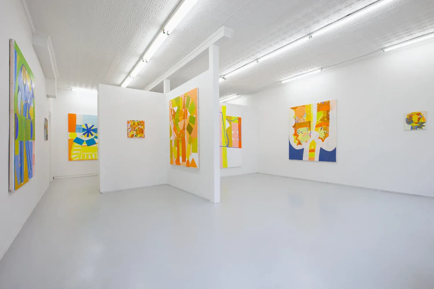 Bright white gallery interior with colorful abstract paintings featuring yellow, blue, and green tones hung on walls and white partitions.