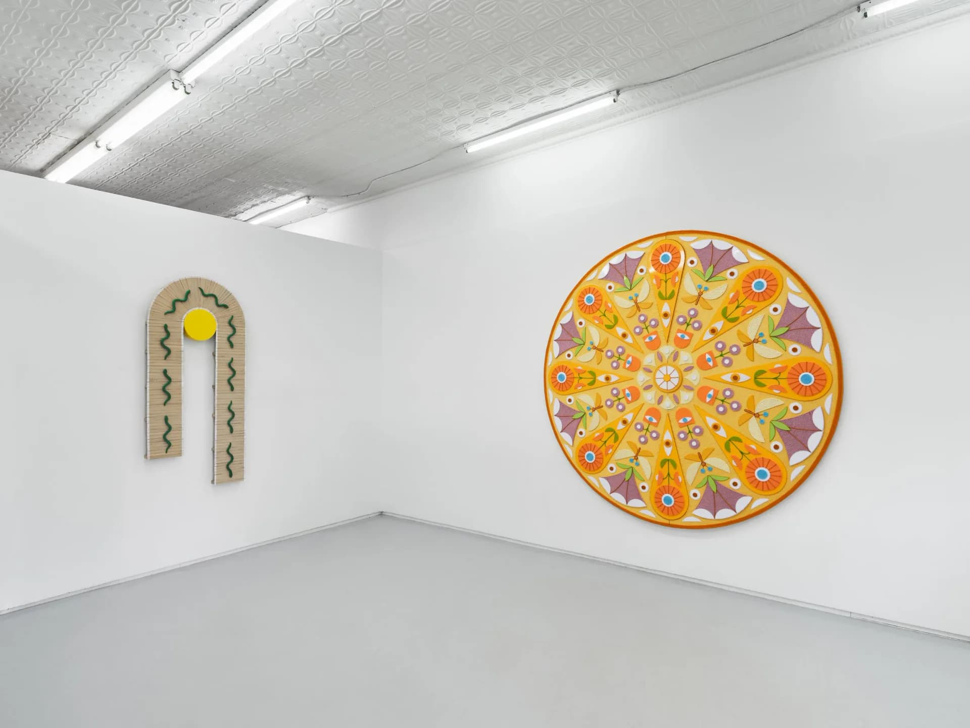 Gallery corner with white walls displaying an arched ornamental piece on left and a large circular yellow floral mandala-like artwork on right.