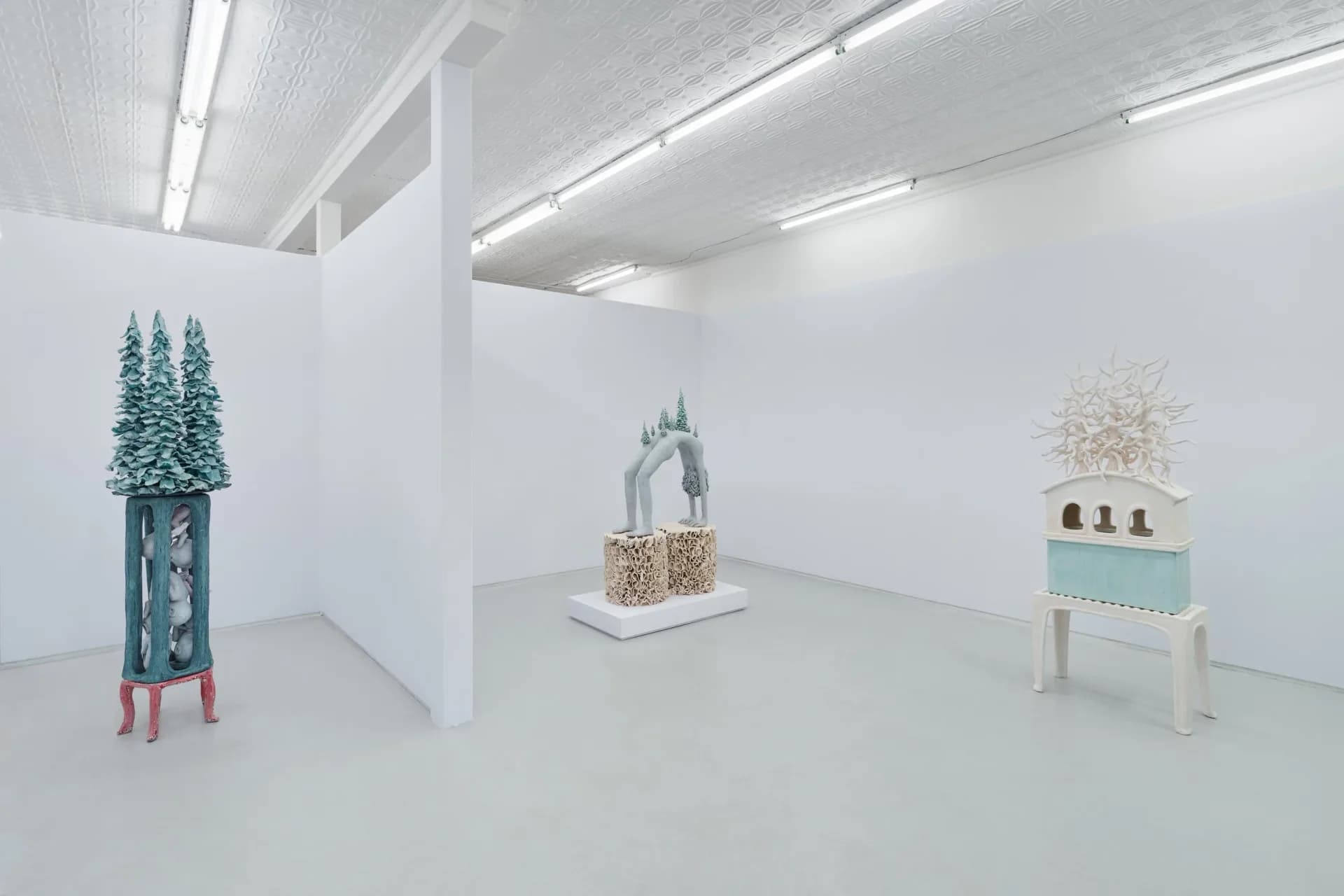 White gallery space featuring three sculptural objects: a tall teal figure with trees, a central cream-colored architectural form, and a light blue cabinet-like structure.