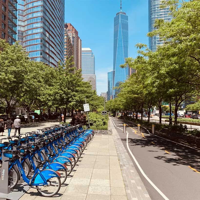 Tree-lined urban street with blue Citi Bikes docked on left, tall Manhattan buildings and One World Trade Center visible in background.