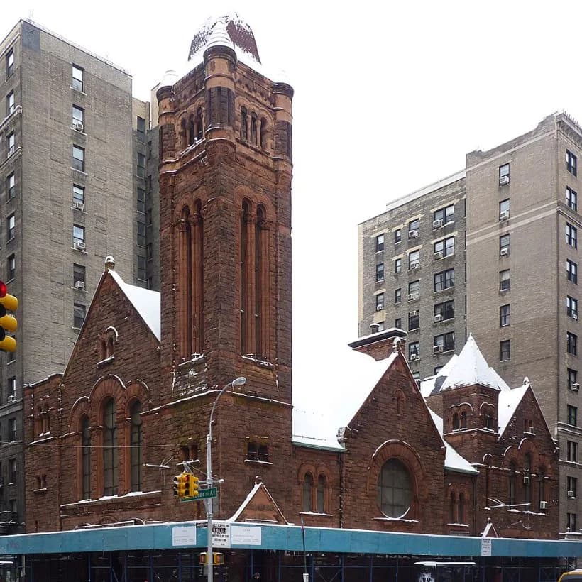Exterior of historic red brick church with ornate tower surrounded by modern gray residential buildings and construction scaffolding.