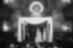Black-and-white image showing a draped canopy structure with two small figures standing beneath ornate arches, evoking a ceremonial or theatrical scene.